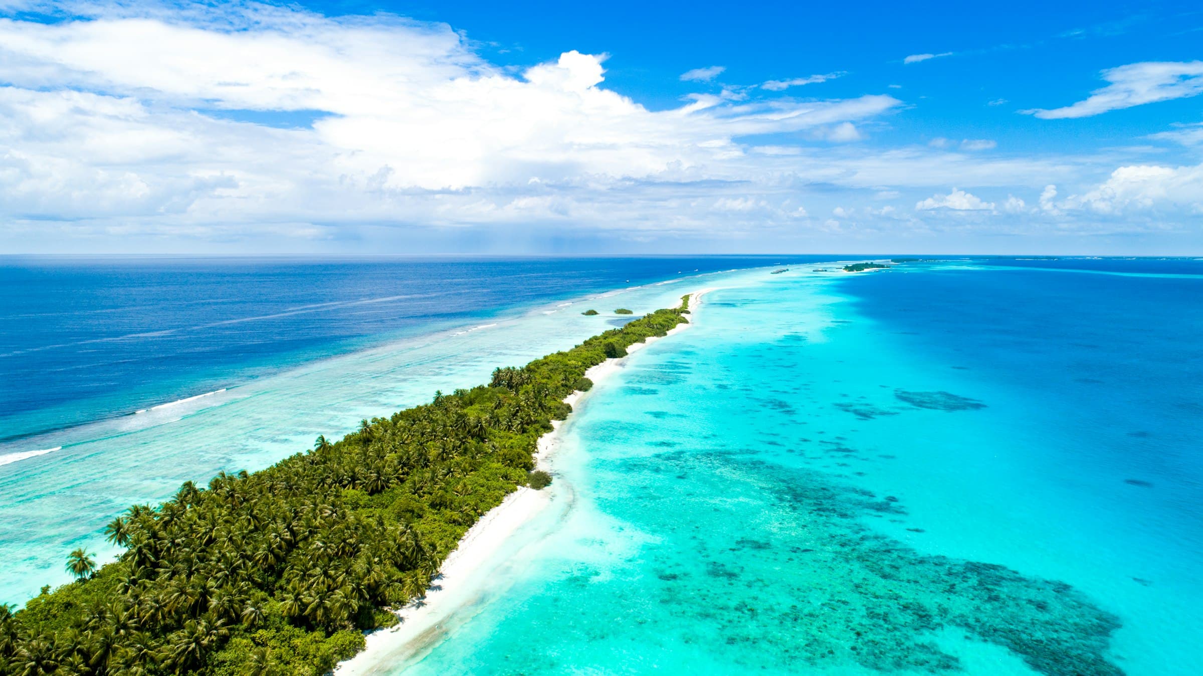 A long, narrow island of palms with a white beach, a turquoise lagoon and reef on one side and deep blue ocean on the other