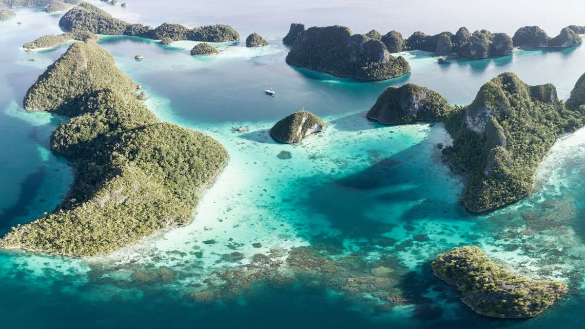 Aerial view of small forested rock islands rising from shallow turquoise water with pale sand and coral patches, and one small boat at anchor