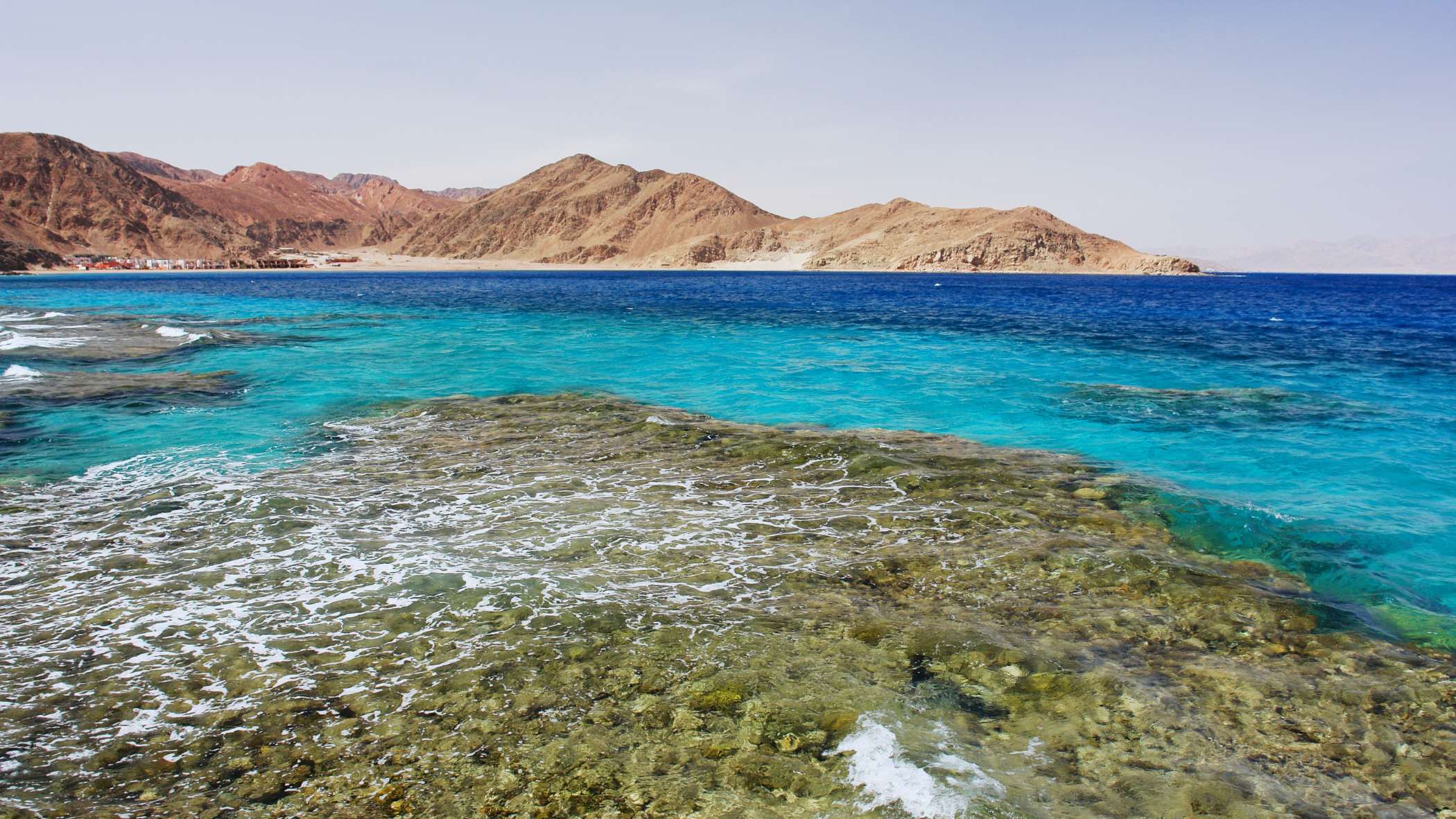 Shallow coral reef flat with small waves breaking over it, turquoise and deep blue water beyond, and bare reddish-brown mountains along the shore