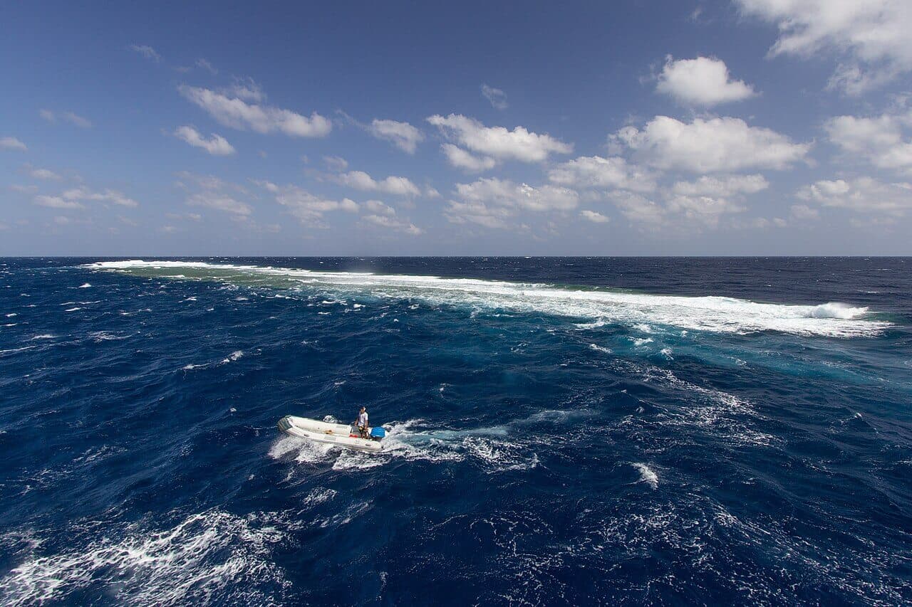 A small white inflatable boat with one person on it, on choppy dark blue water in front of a reef where waves break white