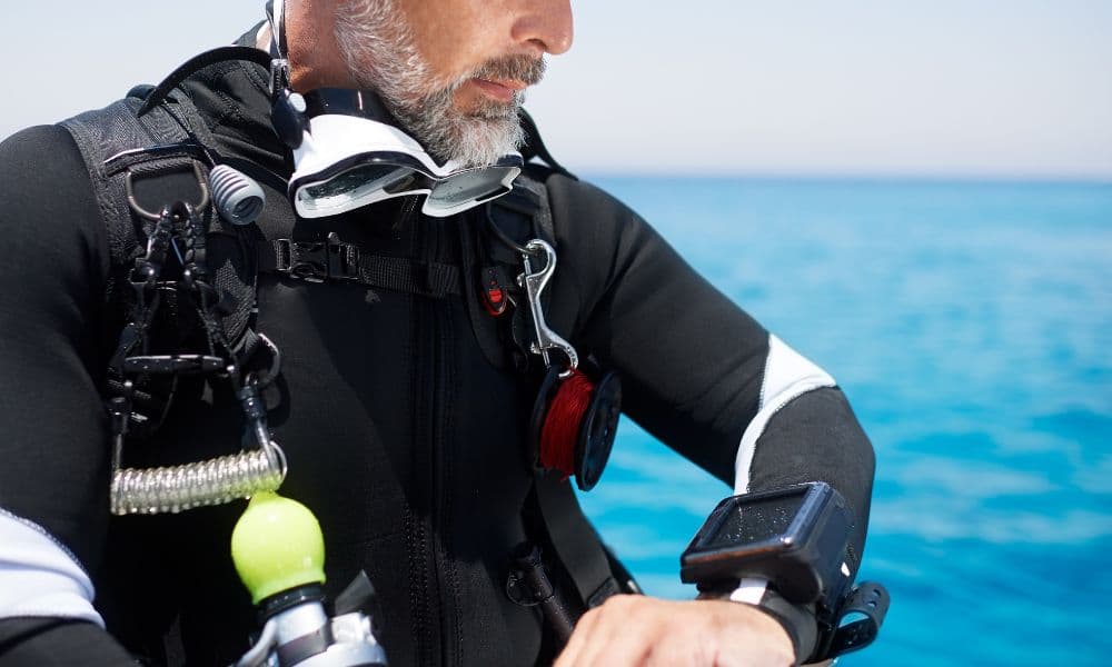 A diver on a boat in a black suit and harness, checking a wrist-mounted dive computer