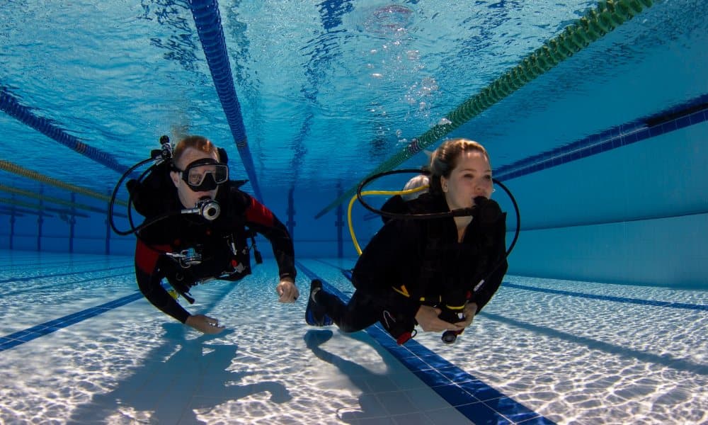 Two divers swim along the bottom of a pool, one of them without a mask