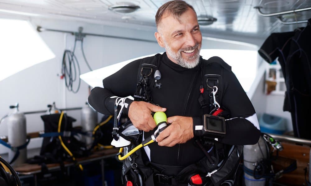 A smiling diver in a wetsuit checks his kit on a dive boat