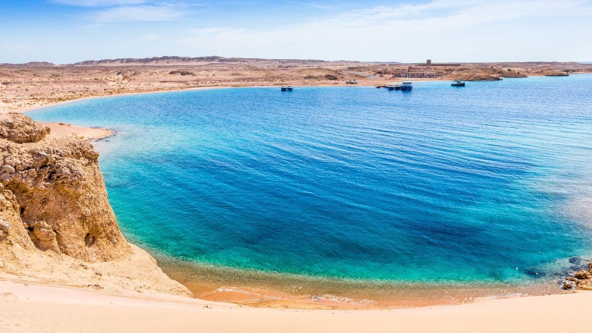 A wide turquoise and deep-blue bay ringed by sandy desert hills and a rocky headland, with a few boats at anchor