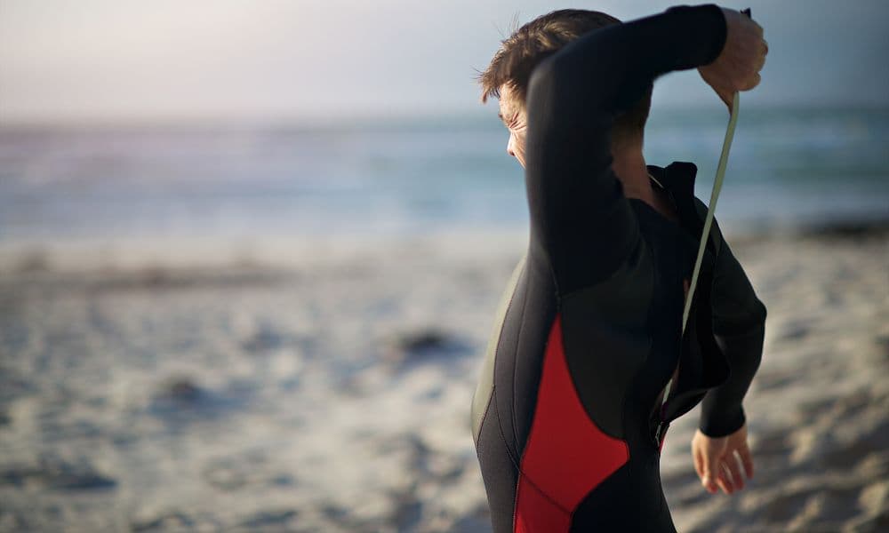 A person pulling on a black and red wetsuit on a beach